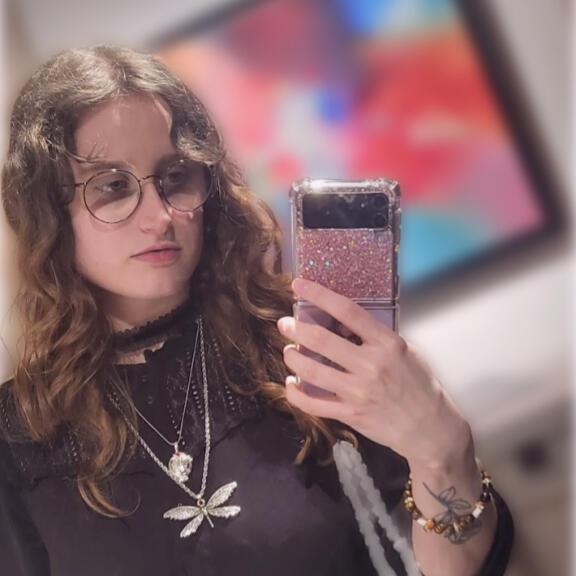 hazyn forsythe taking a mirror selfie with a sparkly pink phone. They have long, wavy auburn hair and wear round glasses, layered silver necklaces (including a dragonfly and circular pendant), a black lace-trimmed dress, and a beaded bracelet. A rose tatto