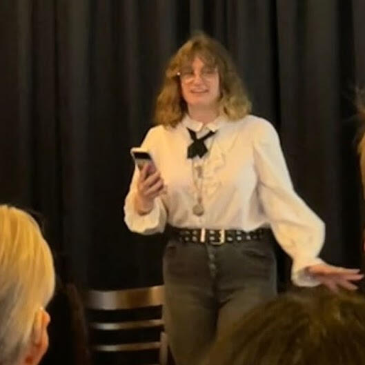 hazyn forsythe reading their piece sealed from Monstera at a 2022 event. They are standing in front of a black curtain, holding a phone, and wearing a white ruffled shirt with a black ribbon tie, layered necklaces, and studded belt. Audience heads are visi