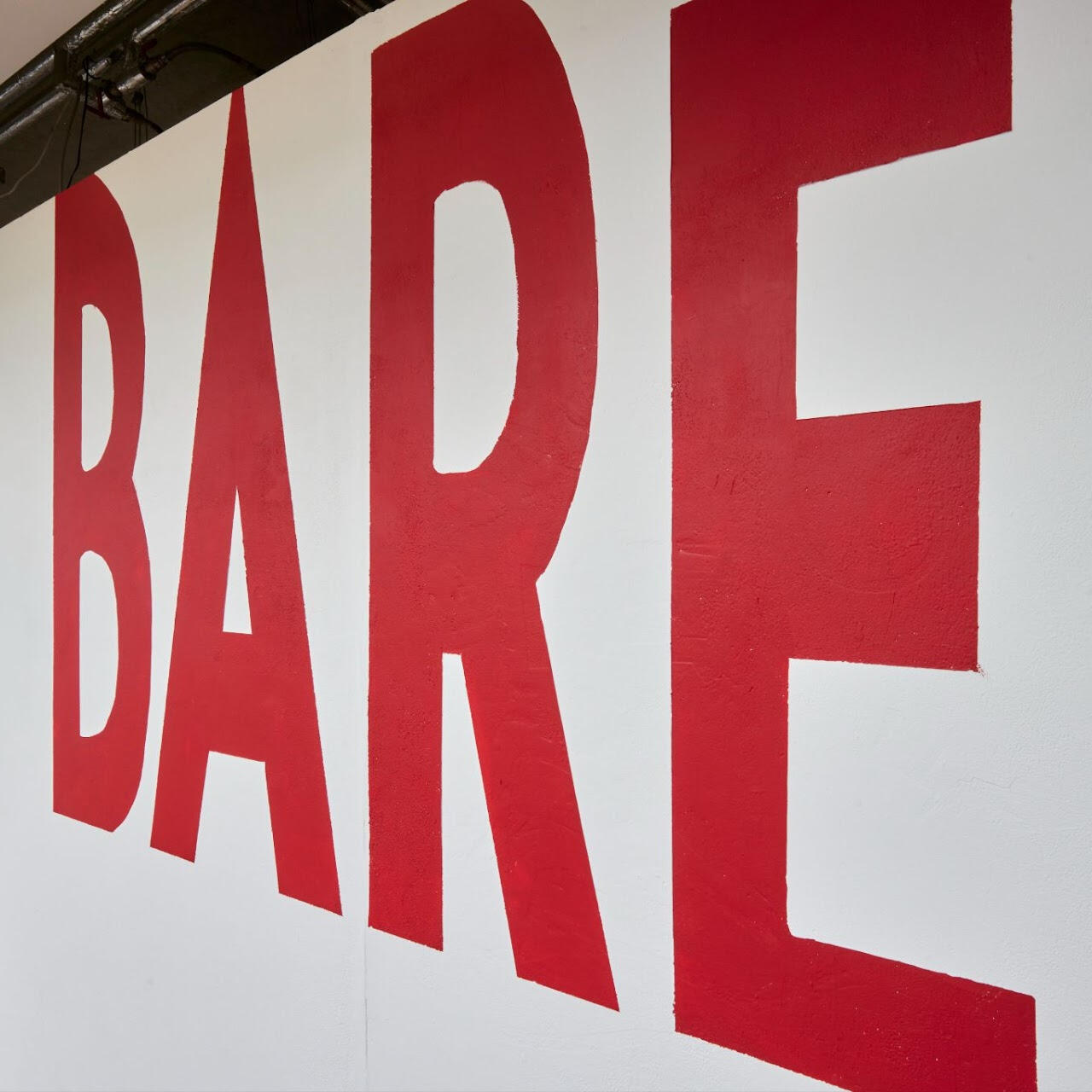 BARE exhibition logo wall, from the BARE website Exhibition title wall for BARE (2023), where and she gave me magic by hazyn forsythe was displayed. The word “BARE” is painted in large, bold red capital letters across a white wall, extending toward the viewer in a dynamic perspective.